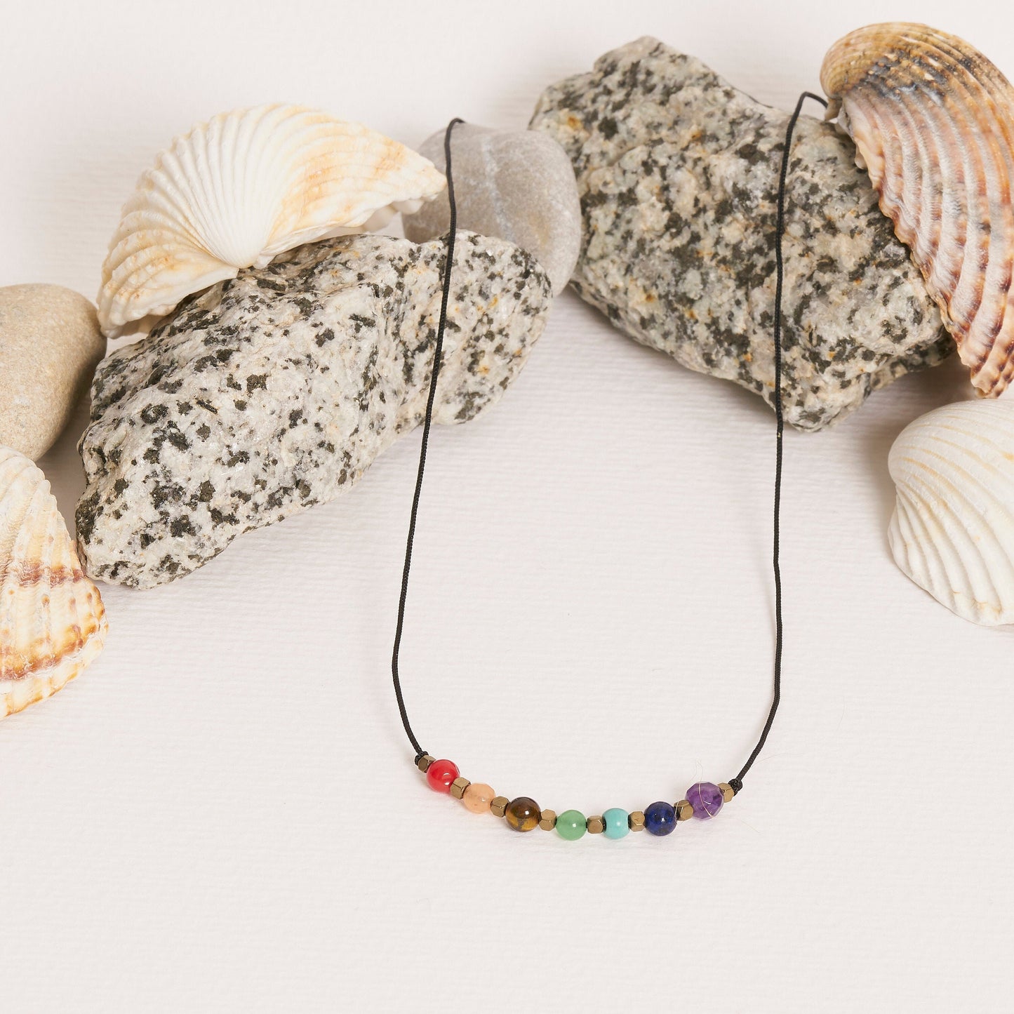 Collar con piedras naturales que representan los 7 chakras, sobre fondo de conchas marinas y rocas decorativas.